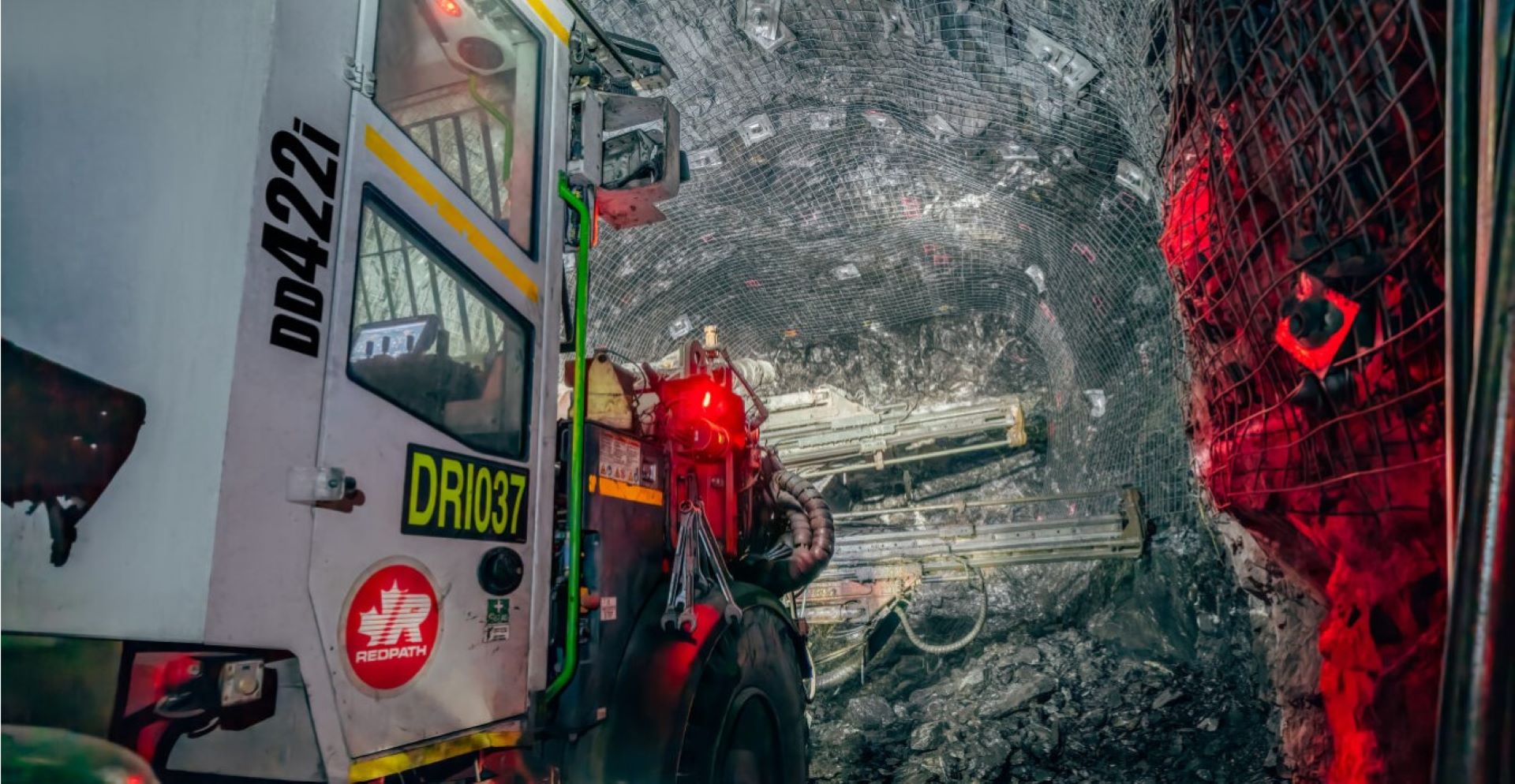 Imagen de un Jumbo Bolter y perforación conductora de apoyo a tierra y superficie mediante la aplicación de malla de alambre en las paredes y el techo de un túnel subterráneo.